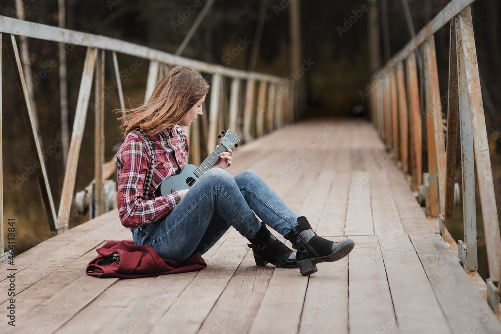 Young woman sitting and playing ukulele guitar on bridge in autumn forest Young woman sitting and playing ukulele guitar on bridge in autumn forest