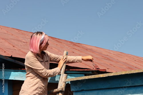 A girl paints an old metal roof with a roller and a brush while standing on a wooden staircase