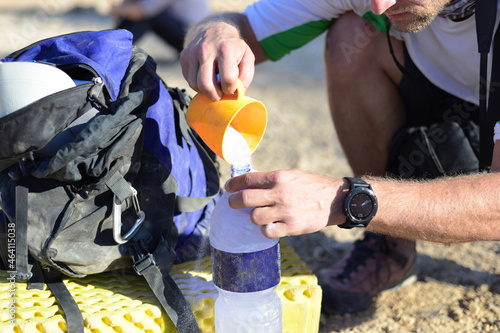 Adventure man pouring Hydration Electrolyte Powder in water bottle. A backpack and a bottle of water with powdered electrolyte on desert mountains ground. man traveler preparing isotonic drink  photo