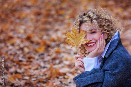 Young beautiful girl smiles and holds a yellow maple leaf in her hand. The girl has curly hair. Autumn Park. Copy space. Blurred background.