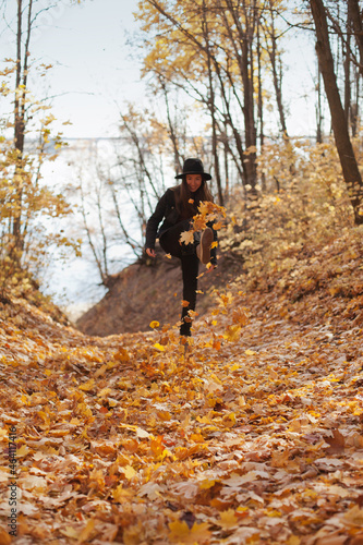 Young woman in black hat having fun in autumn park, throwing fallen autumn leaves in the air