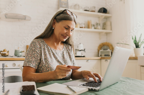 Happy senior woman making online payments of bill using laptop, smiling while shopping with credit card, using application for Internet banking, isolated against kitchen surroundings