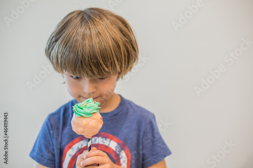 Young Boy Eating a Giant Cupcake Cake Pop with green icing pink chocolate and multi colored Sprinkles at a Birthday Party in front of a wall.