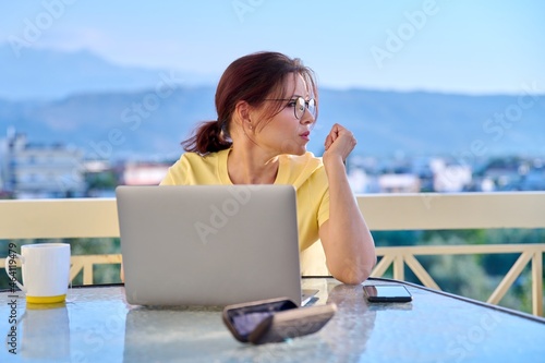 Serious middle aged woman with laptop at table on home outdoor balcony