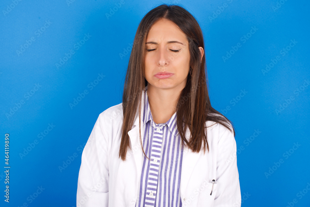 Dismal gloomy rejected young european doctor girl wearing white medical gown on white background has problems and difficulties, curves lower lip and closes eyes in despair, being in depression Dismal gloomy rejected young european doctor girl wearing white medical gown on white background has problems and difficulties, curves lower lip and closes eyes in despair, being in depression