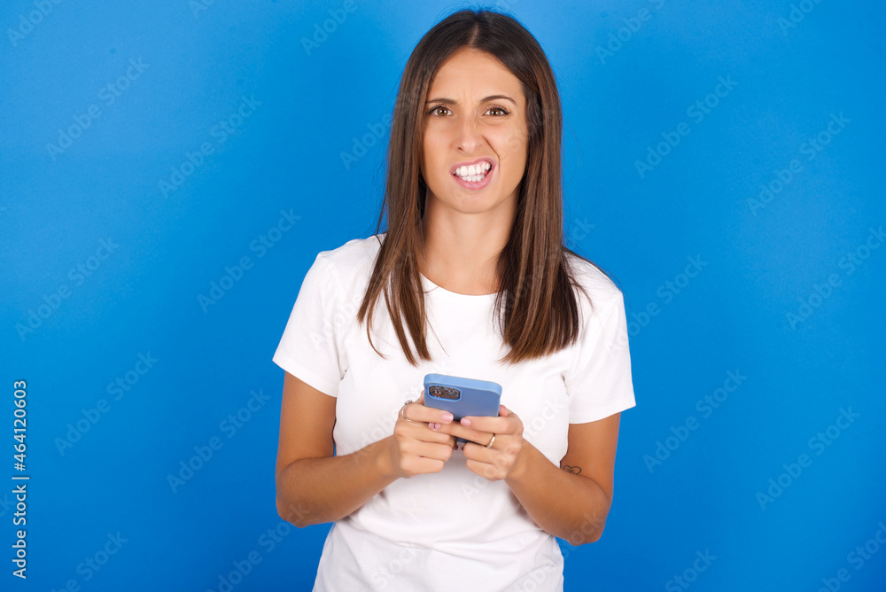 Photo of astonished crazy young european girl wearing white t-shirt on white background hold smartphone dislike feedback concept Photo of astonished crazy young european girl wearing white t-shirt on white background hold smartphone dislike feedback concept
