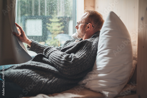 Portrait of thinking sad Middle-aged man in eyeglasses dressed open cardigan lying on cozy bed next to window looking out street through raindrop glass. Mental health and autumn weather concept image photo