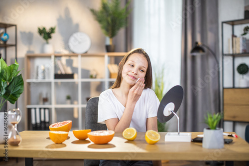 Pretty female teenger sitting at table while cleaning face with cotton pad and looking at mirror. Happy young woman taking care of her healthy pure skin at home. photo