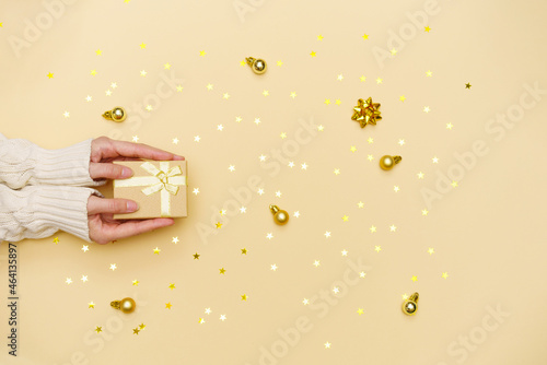 Female hands holding merry christmas gift box on yellow decorated holiday background, woman preparing surprise, new year winter merry holiday golden confetti from stars, real holiday concept, top view © Екатерина Переславце