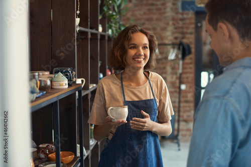 Smiling attractive clerk in apron standing at shelf and selling ceramic cup to man in pottery store photo