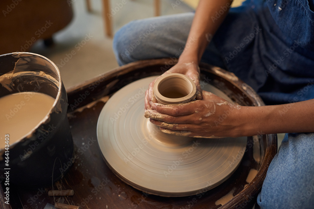 Fototapeta premium Close-up of unrecognizable potter sitting at pottery wheel and creating ceramic pot in workshop