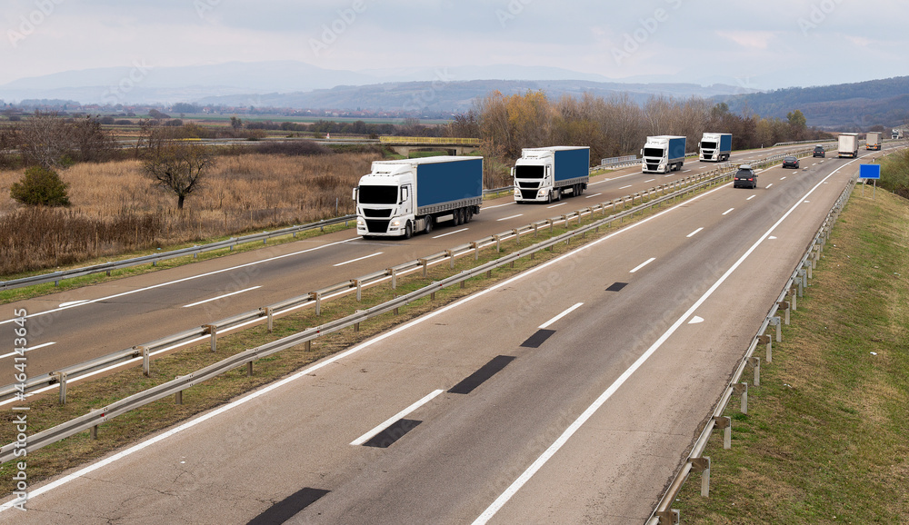 Highway transportation scene with Caravan or Convoy of Blue transportation trucks in line on a rural highway under a dramatic sky