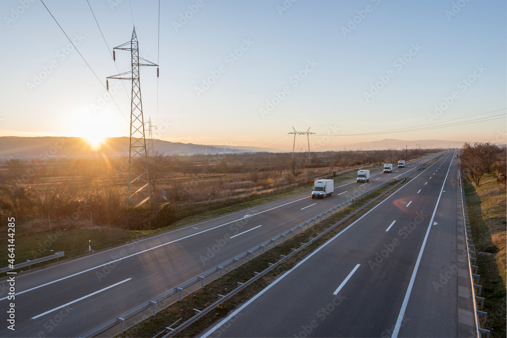 Caravan or convoy of trucks in line on a country highway. Convoy of Big Transportation trucks departing on a country highway under a sunset sky. 