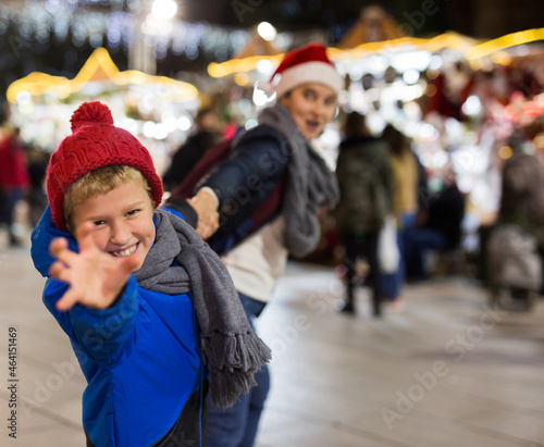 Preteen boy pulling his mom hand, demanding to buy something for him at outdoor Christmas market
