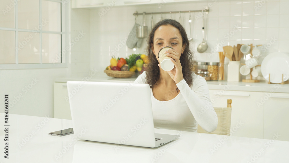 Relax happy latin young businesswoman. People drinking a cup of coffee, using a computer laptop notebook technology device, working online in kitchen at home. Lifestyle