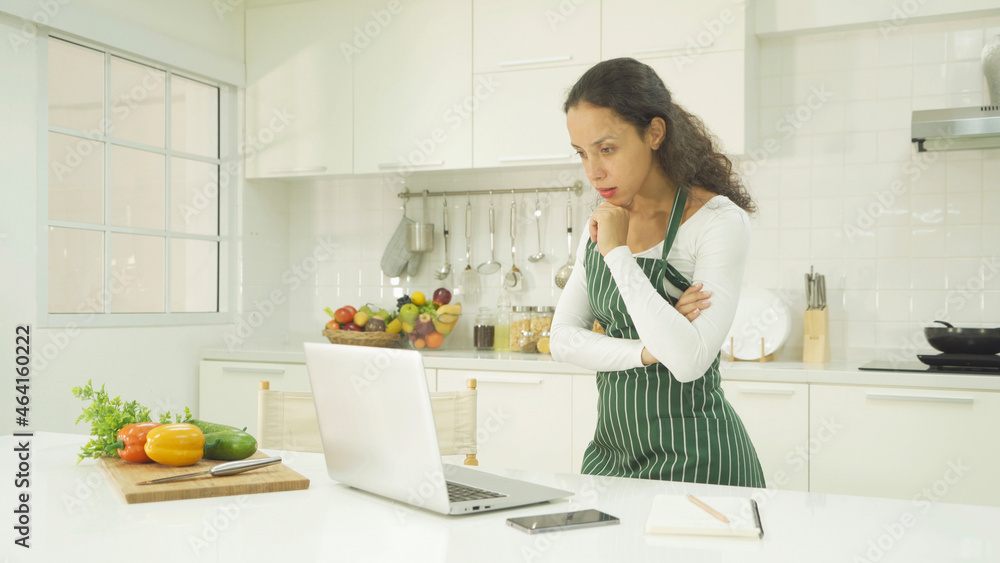 Portrait of happy smiling latin woman using a computer laptop notebook technology device, cooking food, working online at home in kitchen. People lifestyle. Household