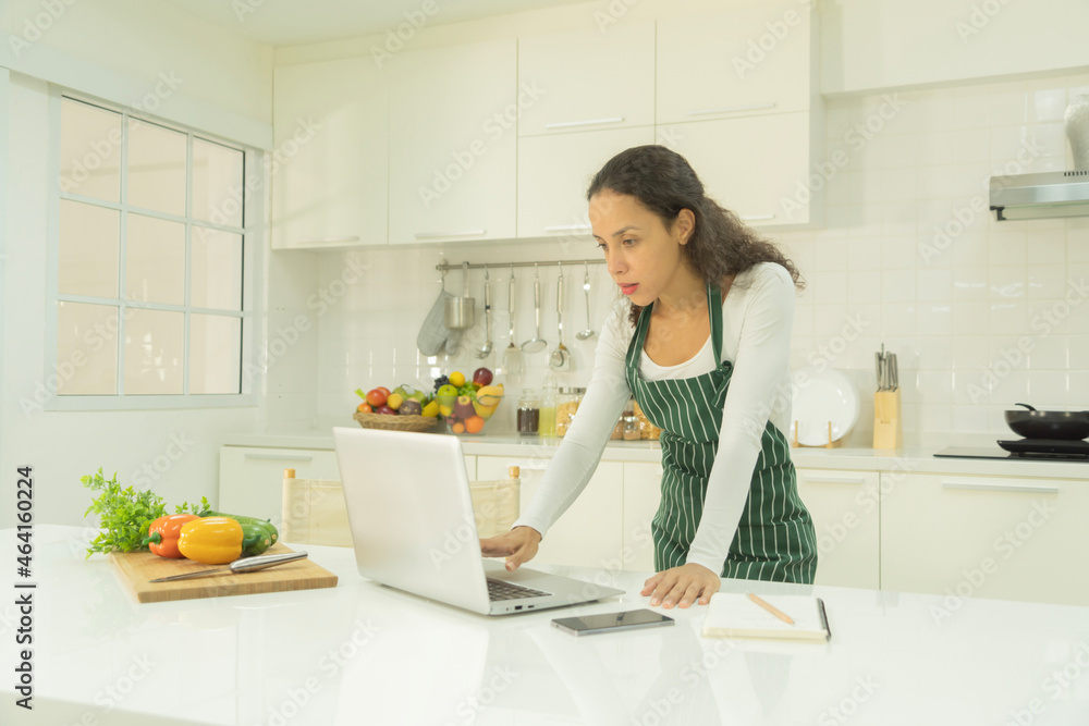 Portrait of happy smiling latin woman using a computer laptop notebook technology device, cooking food, working online at home in kitchen. People lifestyle. Household Portrait of happy smiling latin woman using a computer laptop notebook technology device, cooking food, working online at home in kitchen. People lifestyle. Household
