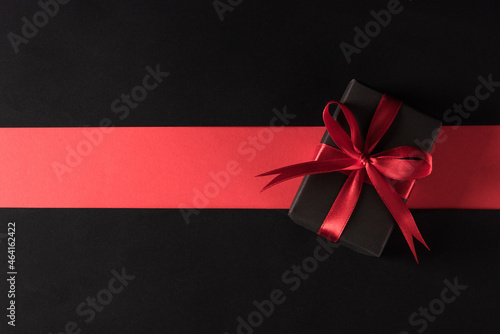Black Friday sale shopping and Boxing Day concept, Gift box wrapped black paper and red bow ribbon present, studio shot isolated on red and dark background