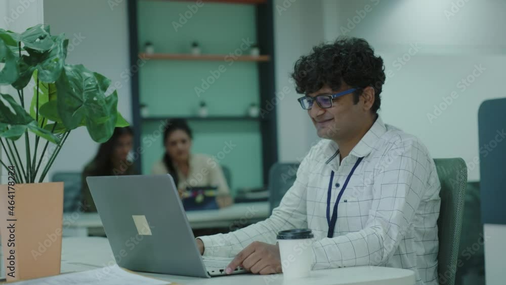 Happy young indian businessman sit on desk using laptop thinking and typing on keyboard work from office.Asian handsome man in a office sits in a suit, drinks coffee and looks at a laptop. Happy young indian businessman sit on desk using laptop thinking and typing on keyboard work from office.Asian handsome man in a office sits in a suit, drinks coffee and looks at a laptop.