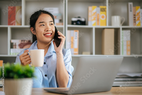 Cheerful young pretty woman use laptop computer and smartphone to work or learning online, hold coffee mug in hand and smile with happy during quarantine covid-19 self isolation at home