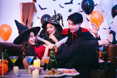 Three Happy friends sitting on sofa and wearing Halloween costumes having fun with party at nightclub or home. Group of young Asian people enjoying Halloween party with many drink and sweets.