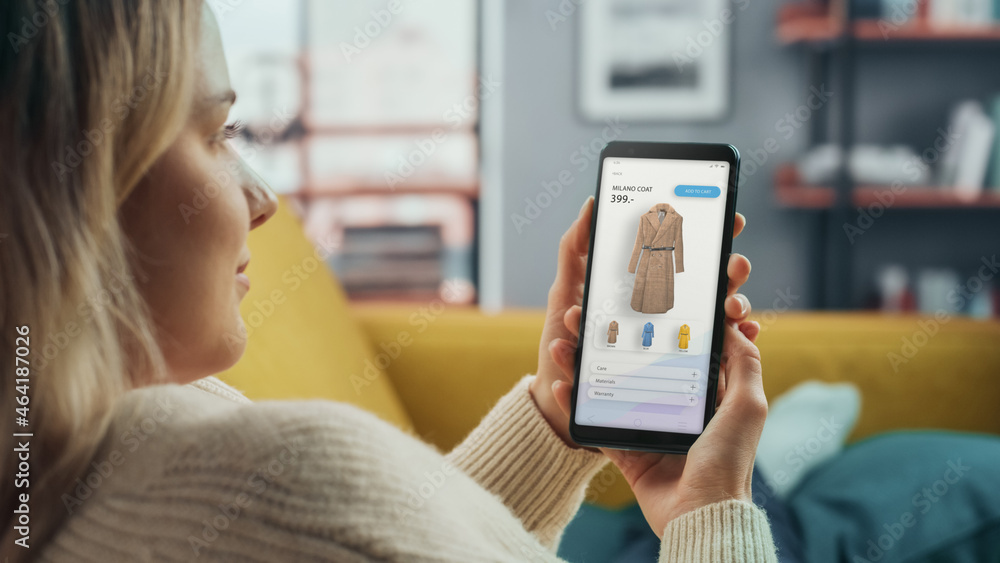 Beautiful Caucasian Female is Using Smartphone with Clothing Online Web Store to Choose and Buy Coat from New Collection. Female Surfing the Net and Lying on Couch Sofa at Home Living Room. Beautiful Caucasian Female is Using Smartphone with Clothing Online Web Store to Choose and Buy Coat from New Collection. Female Surfing the Net and Lying on Couch Sofa at Home Living Room.
