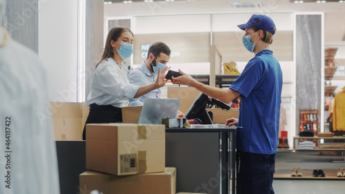 Clothing Store Checkout Counter: Retail Sales Managers wearing Protective Face Masks Give Package to Courier for Online Order Delivery. Sustainable, Stylish Designer Brands Available on Internet.