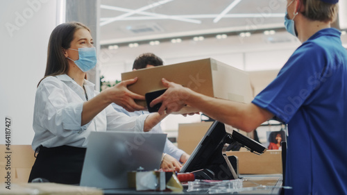 Clothing Store Checkout Cashier Counter: Female and Male Retail Sales Managers wearing Protective Face Masks Give Package to Online Order Delivery Courier. Designer Brands Available to Buy on Internet