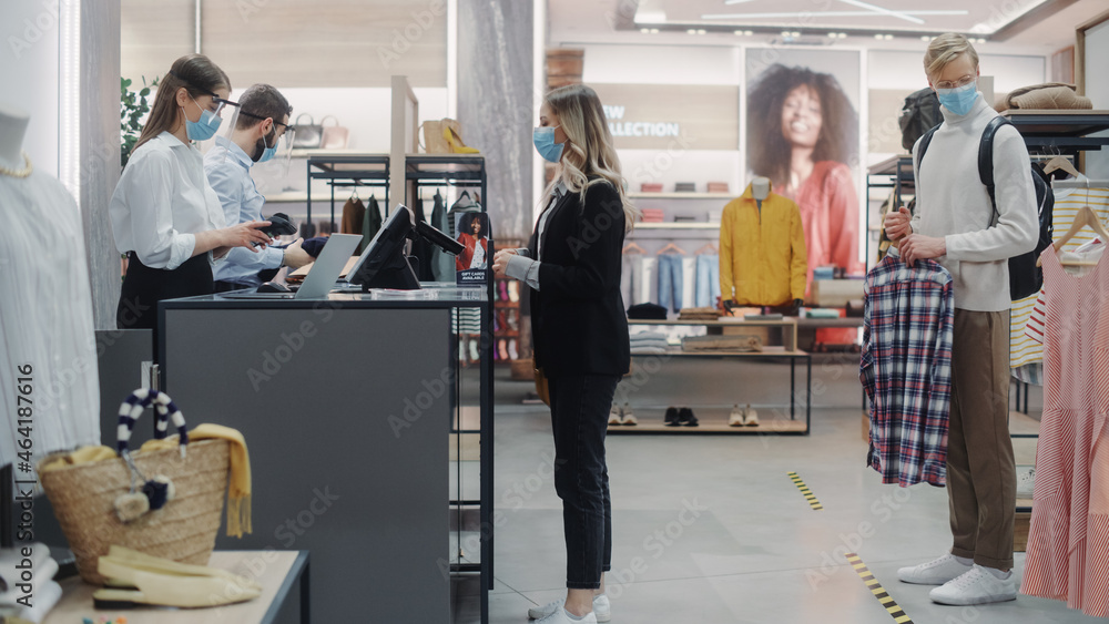 Fototapeta premium Clothing Store: Young Woman At Counter Buys Clothes from Friendly Retail Sales assistant, Paying with Contactless Credit Card. Everybody Wearing Face Masks. Trendy Fashion Shop with of Designer Brands