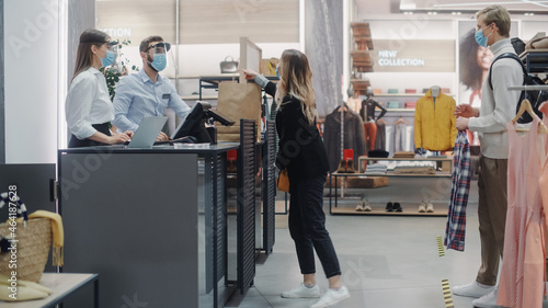 Clothing Store: Young Woman At Counter Buys Clothes from Friendly Retail Sales assistant, Receiving Bag with Purchases. Everybody Wearing Face Masks. Trendy Fashion Shop with of Designer Brands