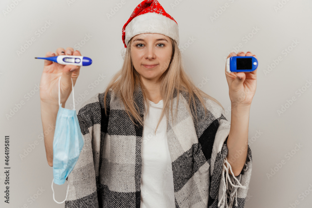 Woman in santa hat, wrap and protective medical mask holds thermometer and puls oximetr Woman in santa hat, wrap and protective medical mask holds thermometer and puls oximetr