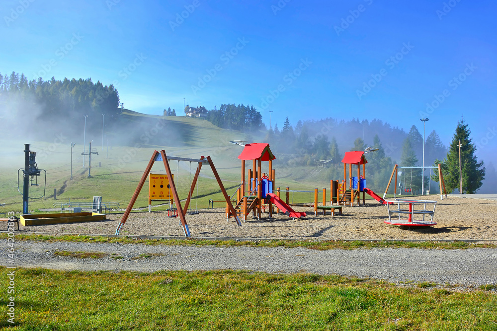 Fototapeta premium Vast green grassland and children's play area,let visitor take a deep breath in the embrace of nature. Foggy mountains on the background, Czorsztyn-Ski, Poland