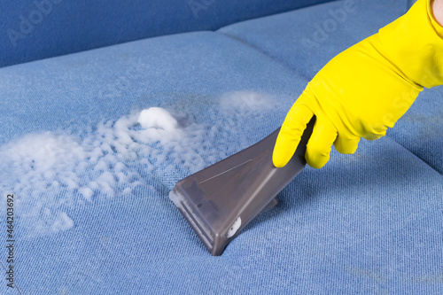 close-up of a hand in yellow gloves cleaning the blue sofa cloth using a washing vacuum cleaner photo
