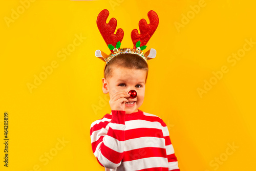 Happy funny toddler boy in red nose and horns reindeer with gift on yellow colored background
