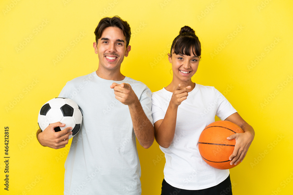 young sport couple playing football and basketball isolated on yellow background points finger at you with a confident expression