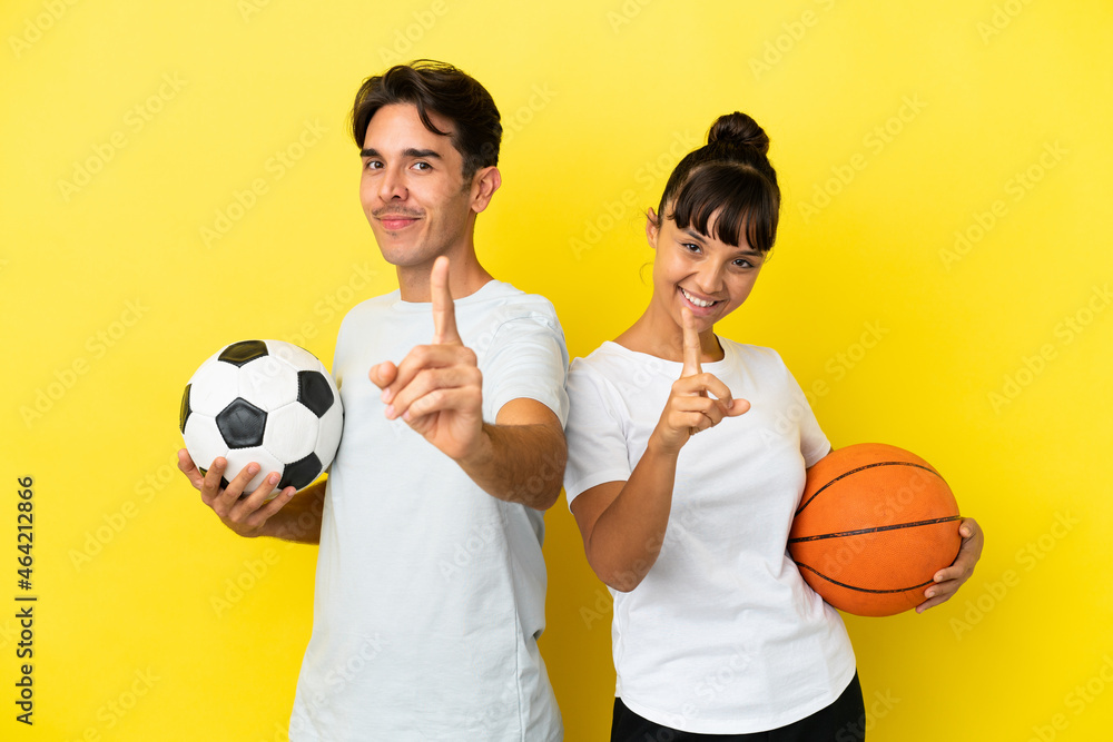 young sport couple playing football and basketball isolated on yellow background showing and lifting a finger