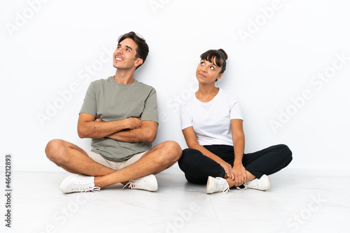 Young mixed race couple sitting on the floor isolated on white background posing with arms at hip and laughing