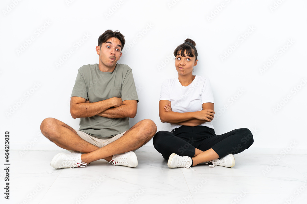Fototapeta premium Young mixed race couple sitting on the floor isolated on white background making doubts gesture while lifting the shoulders