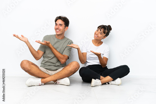 Young mixed race couple sitting on the floor isolated on white background inviting to come with hand. Happy that you came