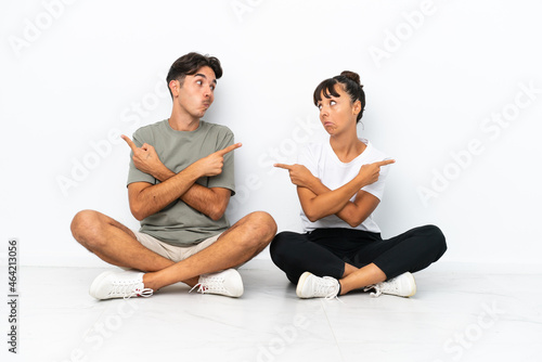 Young mixed race couple sitting on the floor isolated on white background pointing to the laterals having doubts