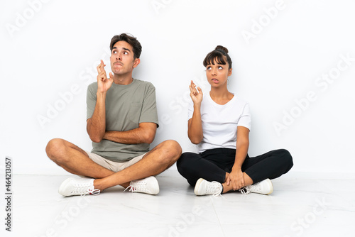 Young mixed race couple sitting on the floor isolated on white background with fingers crossing and wishing the best