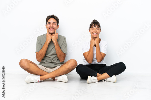 Young mixed race couple sitting on the floor isolated on white background smiling with a happy and pleasant expression