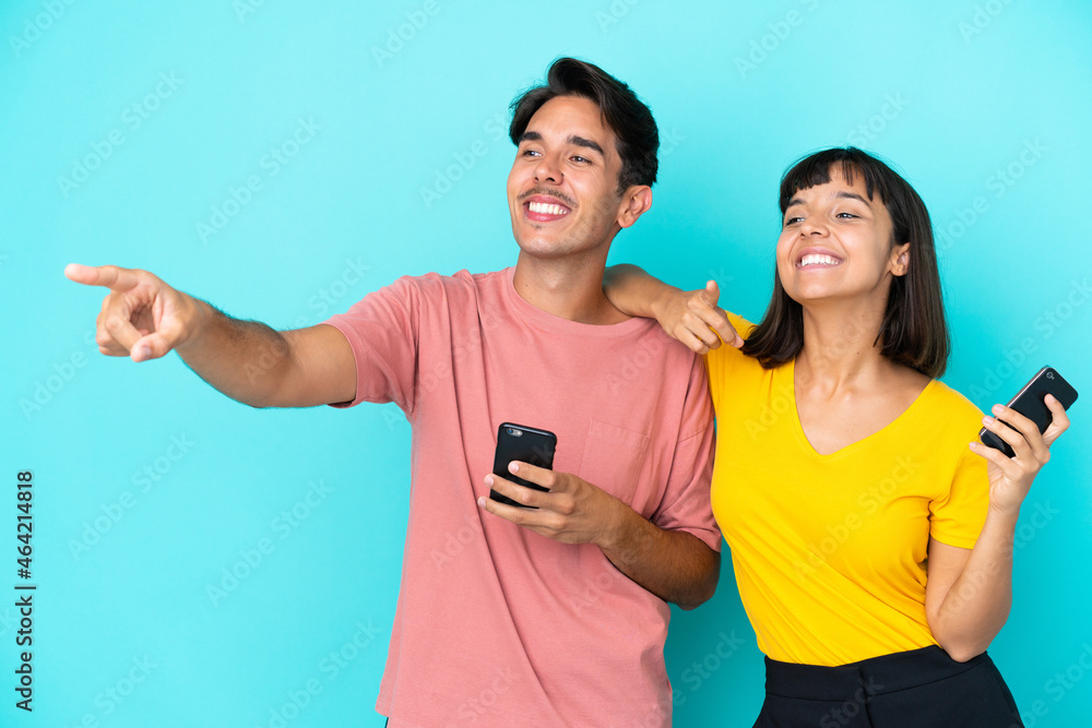 Young mixed race couple holding mobile phone isolated on blue background presenting an idea while looking smiling towards Young mixed race couple holding mobile phone isolated on blue background presenting an idea while looking smiling towards