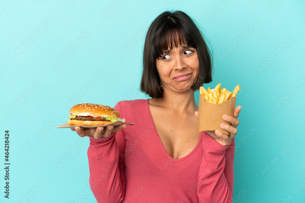 Young brunette woman holding burger and fried chips over isolated blue background