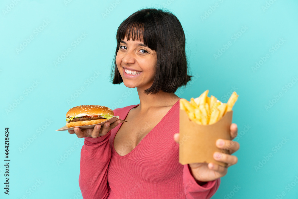 Young brunette woman holding burger and fried chips over isolated blue background Young brunette woman holding burger and fried chips over isolated blue background