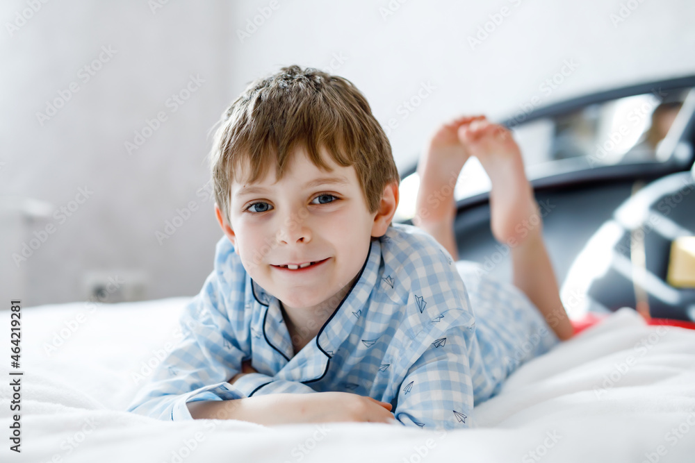 Adorable happy little kid boy after sleeping in his white bed in colorful nightwear. School child celebrating pajama party and looking at the camera. Funny happy child playing and smiling Adorable happy little kid boy after sleeping in his white bed in colorful nightwear. School child celebrating pajama party and looking at the camera. Funny happy child playing and smiling