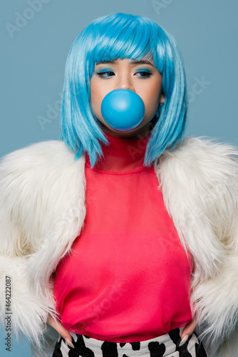 Asian woman in furry jacket blowing bubble gum isolated on blue © LIGHTFIELD STUDIOS