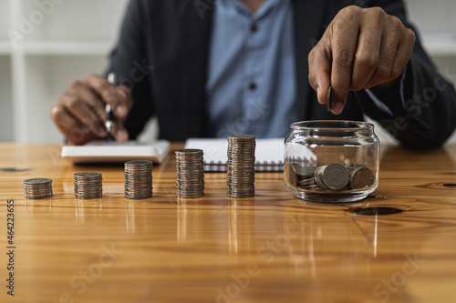 A man is dropping coins into a glass jar with a number of coins inside, on a desk are stacked coins from low to high showing the growth of money. Concept of saving and investing money.