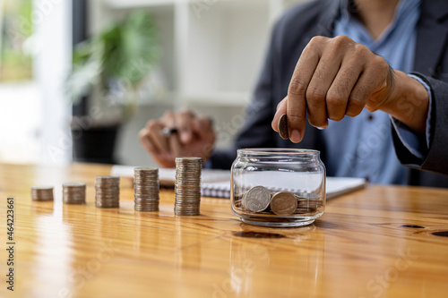 A man is dropping coins into a glass jar with a number of coins inside, on a desk are stacked coins from low to high showing the growth of money. Concept of saving and investing money.