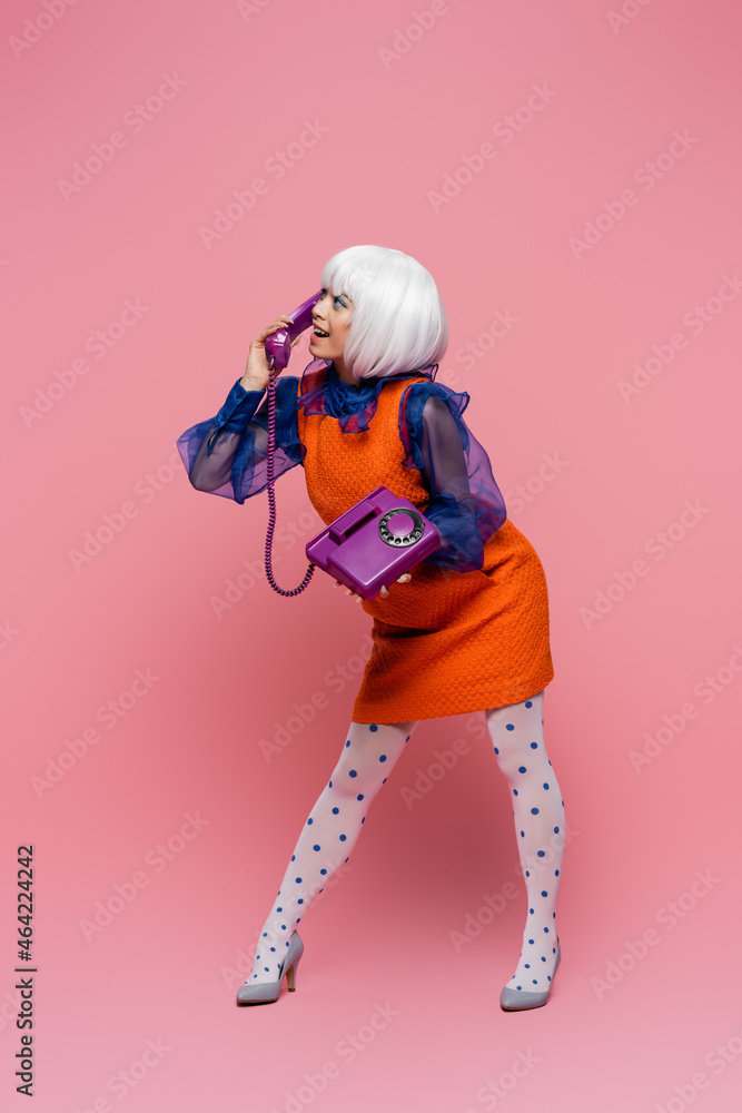Obraz premium Smiling asian woman in vintage dress and white wig talking on telephone on pink background